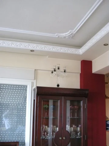Σαλόνι με διαδοχικές λευκές κορνίζες οροφής και διακοσμητική γωνιακή σύνθεση.